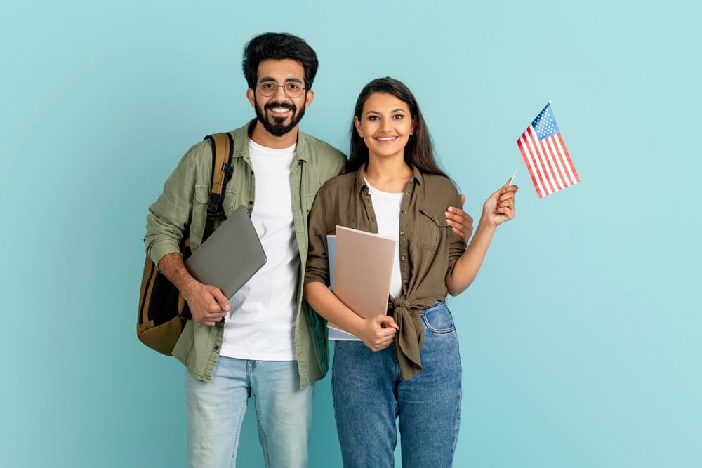Two smiling young adults stand together holding laptops and an American flag against a light blue background, inspired by the achievements of a mandamus lawyer.
