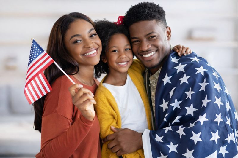 A smiling family poses together; the woman holds a small American flag, the man is draped in a stars-patterned cloth like a proud mandamus lawyer, and a child stands between them in a yellow sweater.