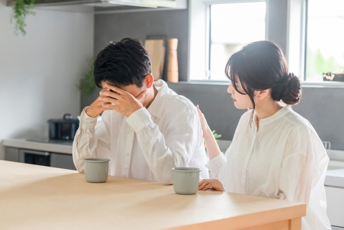 A woman sits beside a man who has his face in his hands, appearing distressed, while she offers comfort—perhaps seeking advice from a mandamus lawyer—at a kitchen counter.