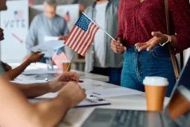 People at a voting station interact, with one person holding a small American flag; papers, coffee cup, laptop, and a mandamus lawyer assisting are visible on the table.