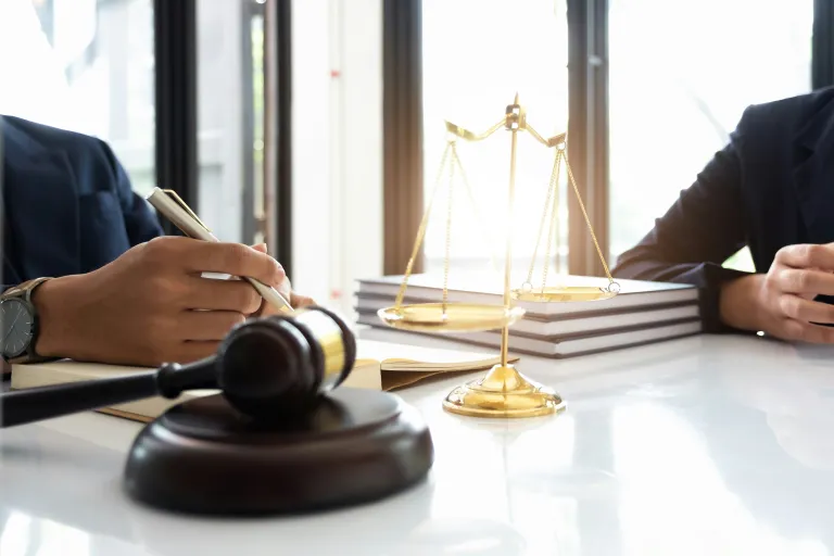 Two people sit at a desk with legal documents, a gavel, and a golden balance scale, as a mandamus lawyer discusses their legal options in this courtroom setting.