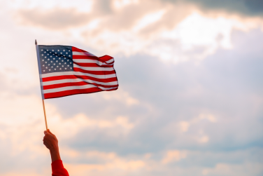 A hand holds up an American flag against a cloudy sky with soft sunlight in the background, symbolizing justice and freedom—core values upheld by any dedicated mandamus lawyer.
