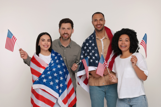 Four people stand together smiling, holding small American flags—perhaps celebrating a mandamus lawyer's recent success. Two are draped in U.S. flag patterns, all in casual clothing, against a plain background.