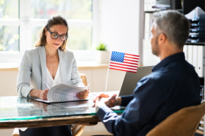 A mandamus lawyer reviews documents while interviewing a man across a desk; a small American flag sits on the desk beside them.