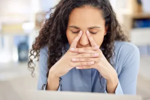 A woman with curly hair sits with her eyes closed, pinching the bridge of her nose and resting her face on her hands, appearing stressed or tired—perhaps awaiting news from her mandamus lawyer.