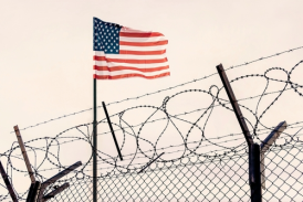 An American flag flies behind a barbed wire fence topped with razor wire under a pale sky, evoking the determination of a mandamus lawyer seeking justice.