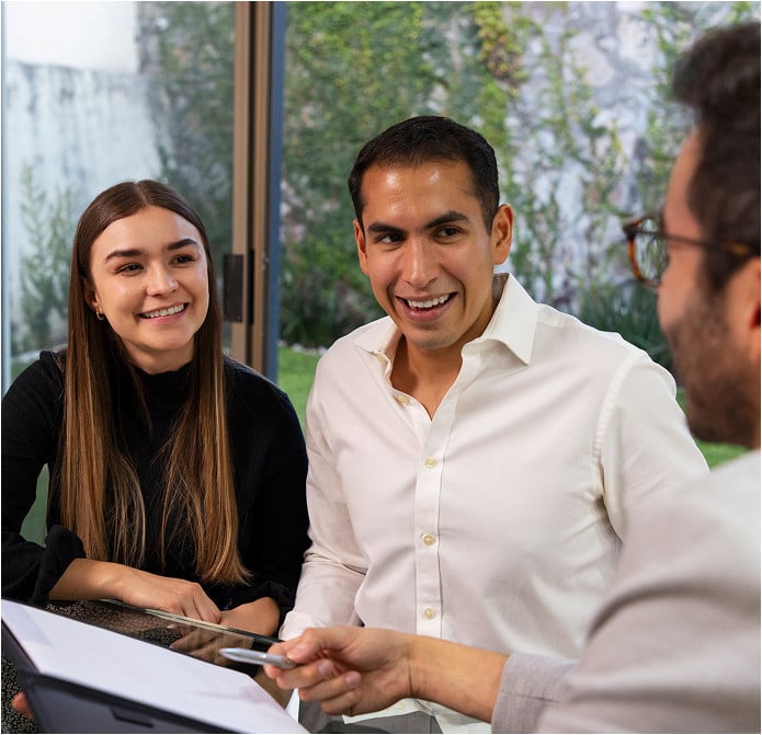 A smiling couple sits at a table with a mandamus lawyer, the man holding a pen and documents, likely discussing paperwork or a contract in a bright indoor setting with a garden view.