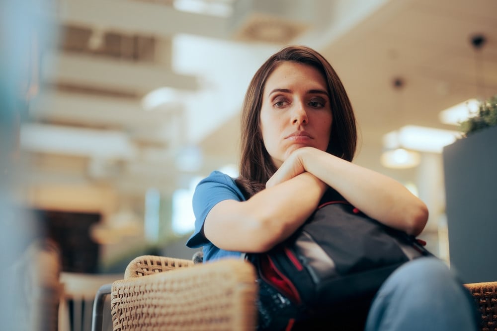 A woman sits indoors with her arms crossed over a backpack, looking pensive and slightly downcast, perhaps awaiting news from her mandamus lawyer.