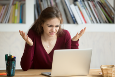 A woman sits at a desk with an open laptop, raising her hands in confusion—perhaps searching for answers from a mandamus lawyer—with books lining the shelf behind her.