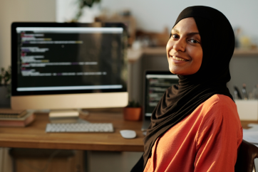 A woman wearing a black hijab and orange top sits at a desk, smiling, with computer screens displaying code in the background—perhaps a mandamus lawyer blending technology with law.