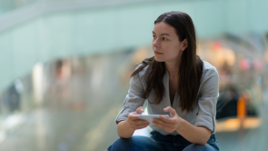 A woman with long brown hair sits indoors, holding a smartphone and looking to the side—perhaps waiting for an update from her mandamus lawyer. The background is blurred.