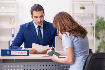 A man in a suit sits at a desk labeled "Visa Denied," reviewing documents, while a woman consults her mandamus lawyer and holds her passport and paperwork across from him.